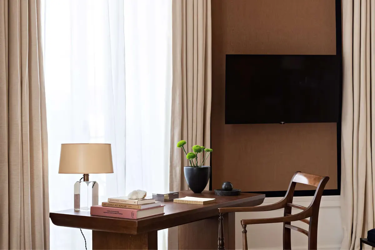 A bedroom with a desk, tan color palette, brown wood furniture, and large window