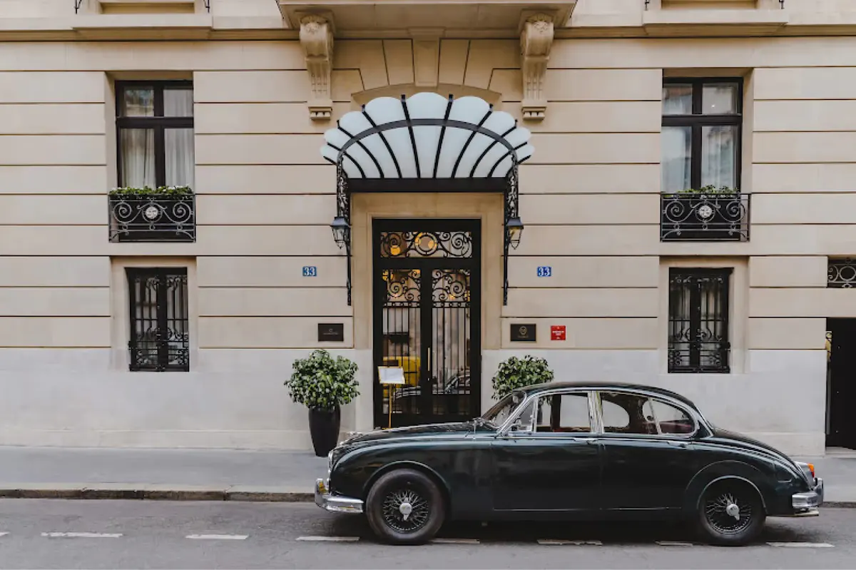 A classical beige facade with arched entry, black-trimmed windows, and a black vintage car parked outside