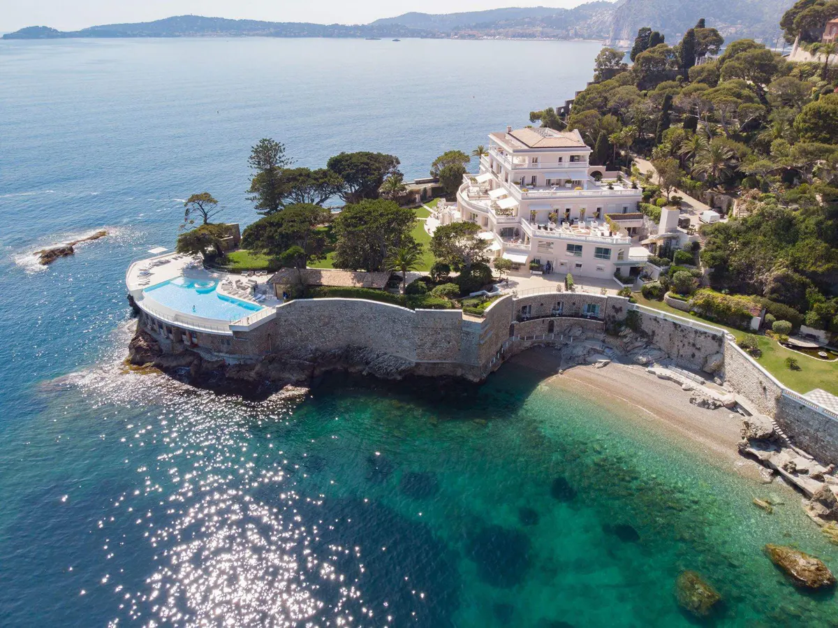 Aerial view of a curved white building on a peninsula with turquoise water and sandy beach