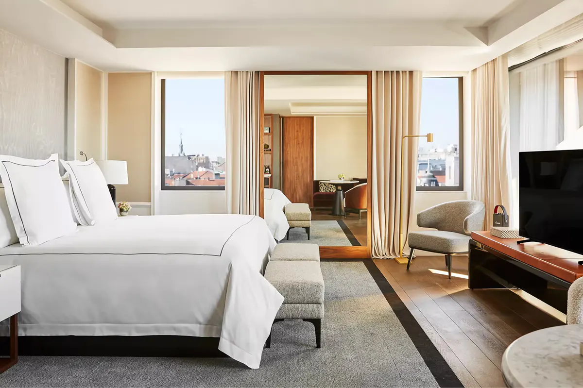 A guest room with white bedding, floor-to-ceiling windows with city views, and warm wood accents