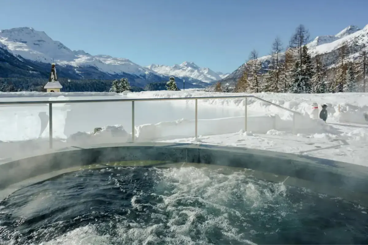 An outdoor hot tub with steaming water, glass railings, and snowy alpine mountain scenery