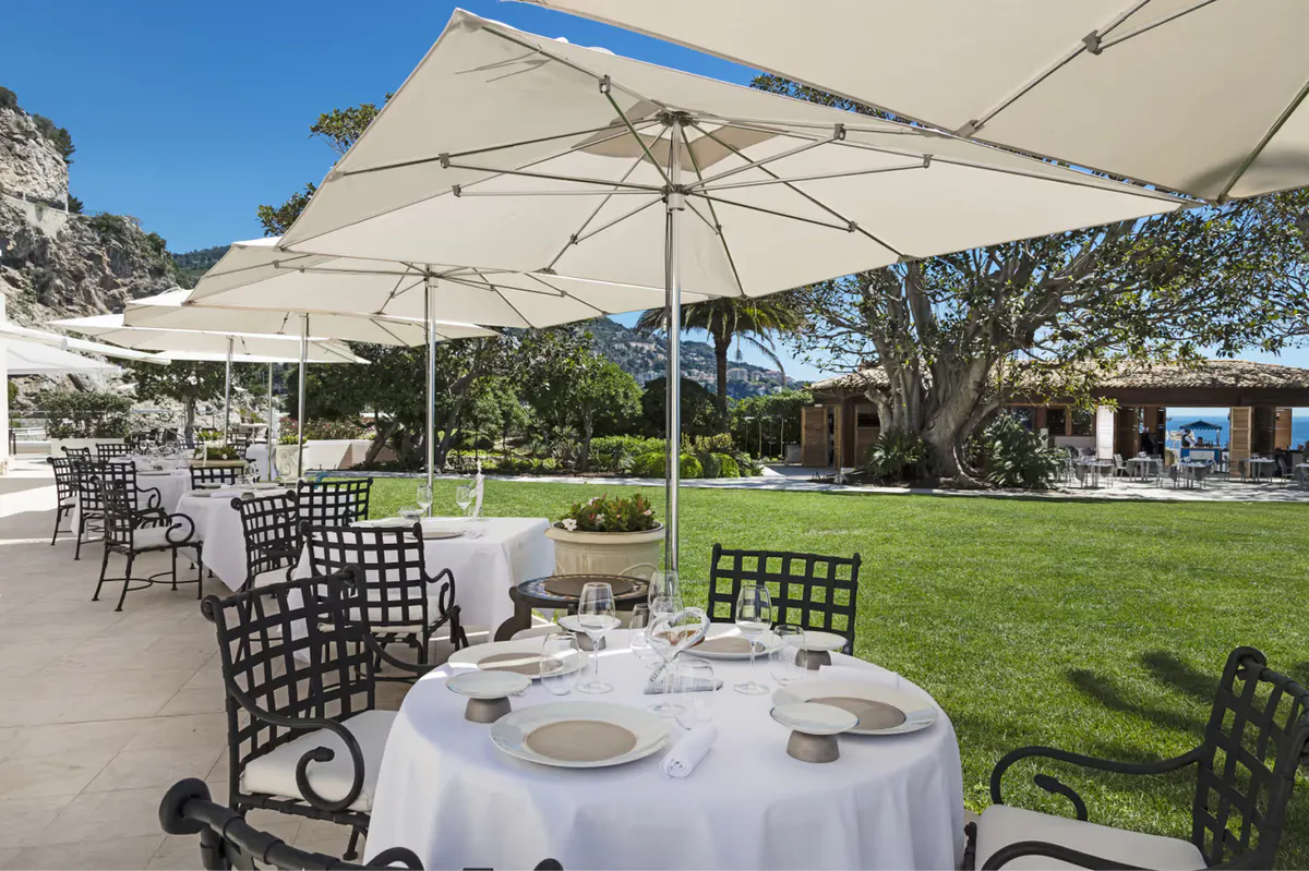 A dining table set with white linens under large umbrellas on a terrace