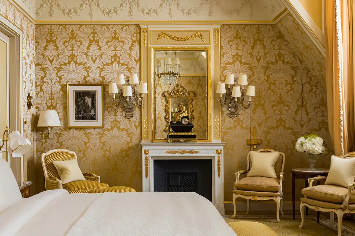 A Belle Époque suite with gold damask walls, cream bedding, fireplace mantel, and gilt-framed mirror