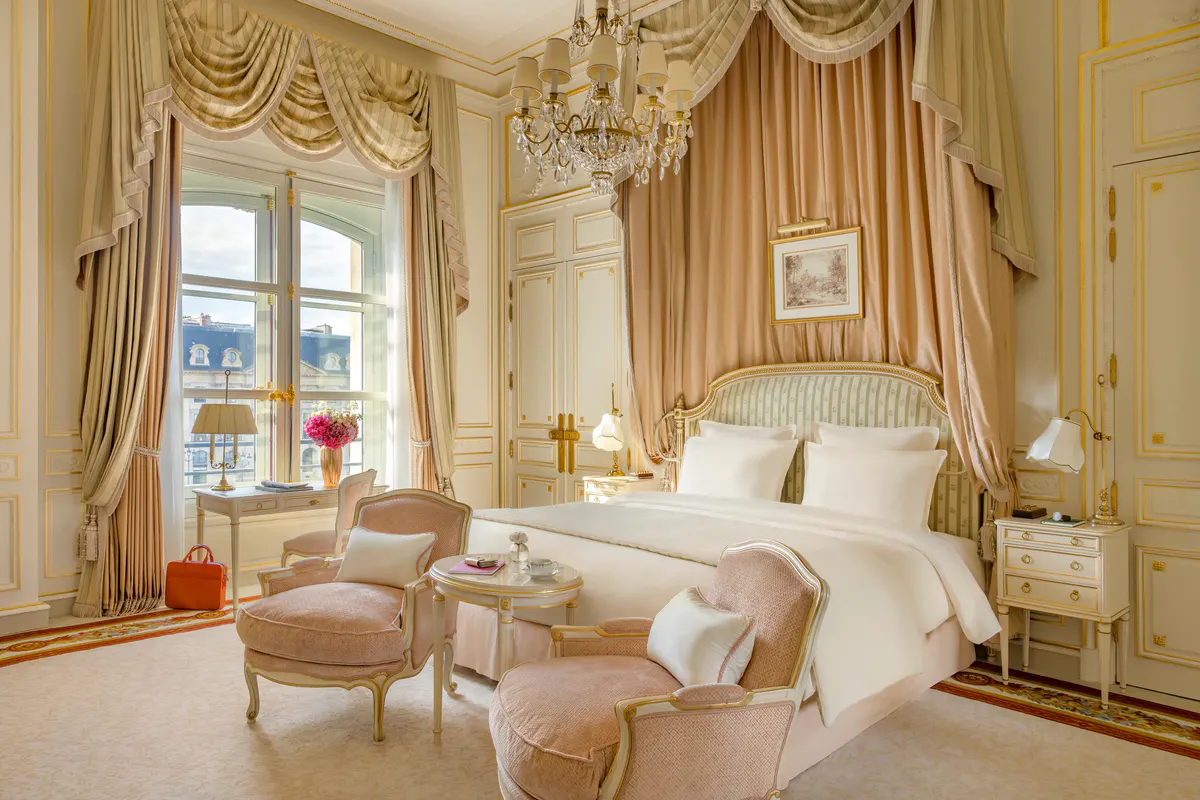 A bedroom with gilt curtain rods, gold drapery swags, cream upholstered seating, and polished hardwood floor