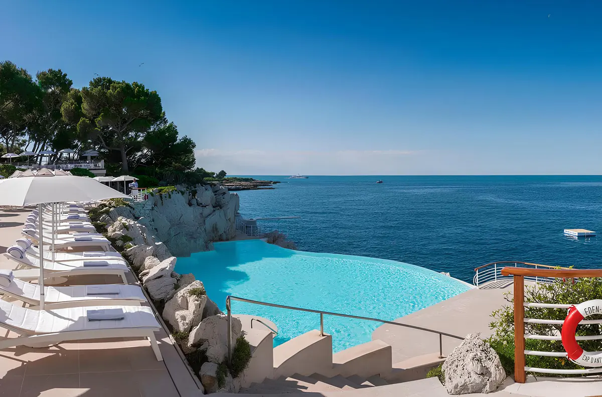 A turquoise infinity pool above the sea, with loungers, bright sun umbrellas, and rocky coastline