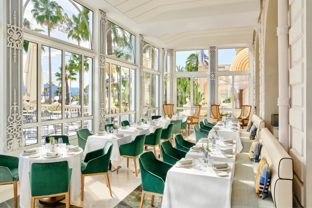 Fine dining room with windows, white tablecloths, green velvet chairs, and ornate ceiling detail