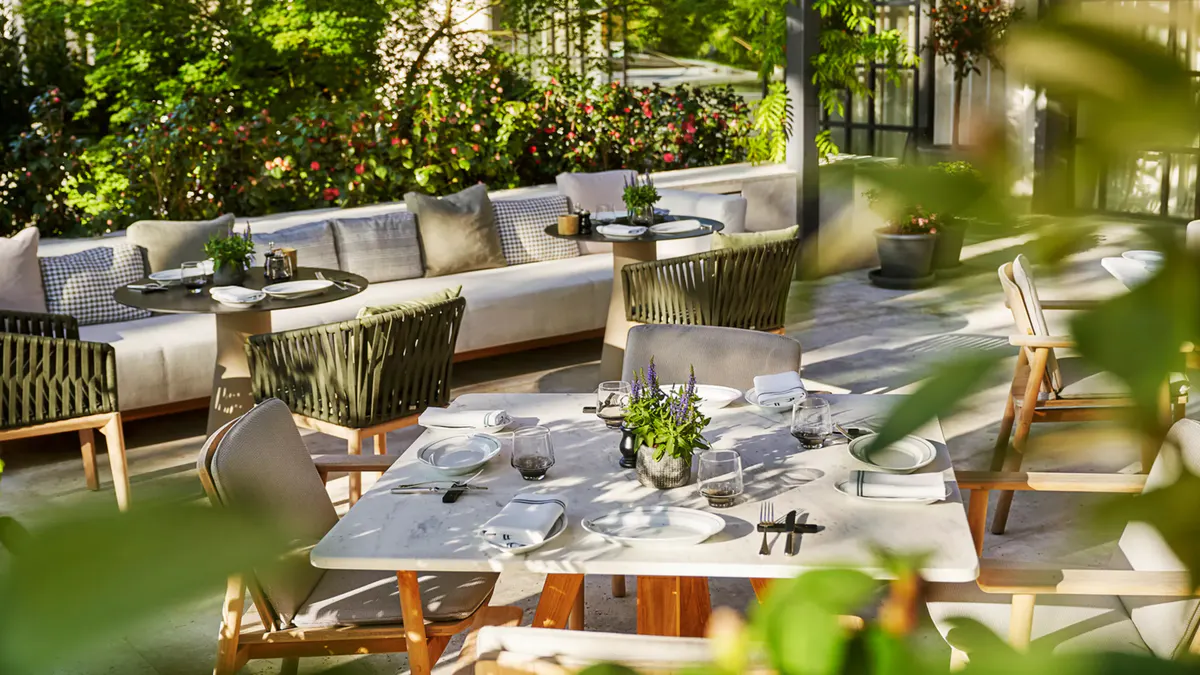 An outdoor courtyard with green seating, tables set for dining, and lush plants in foreground