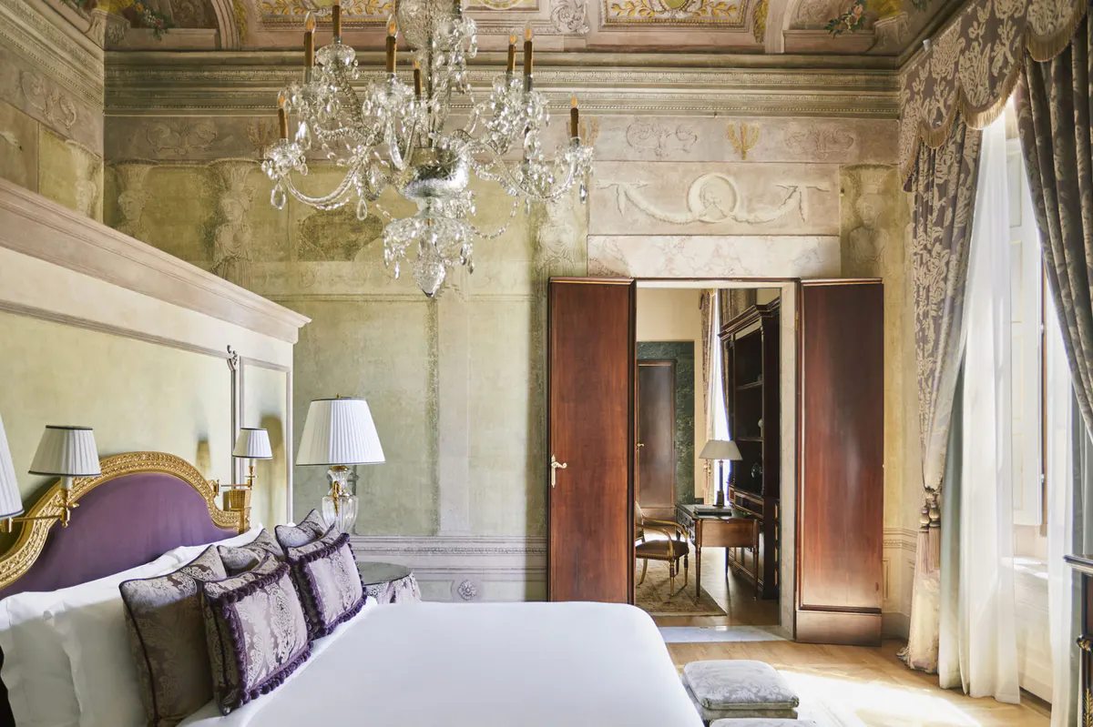 Guest room with pale walls, frescoed ceiling, ornate mirror, and double doors opening to adjacent space