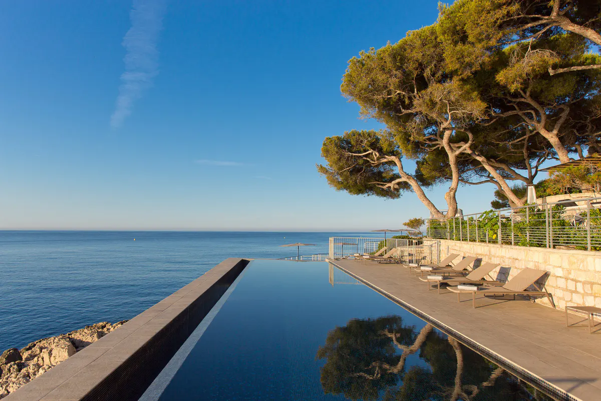 The infinity pool edge with a tall pine tree and Mediterranean Sea in the distance at midday