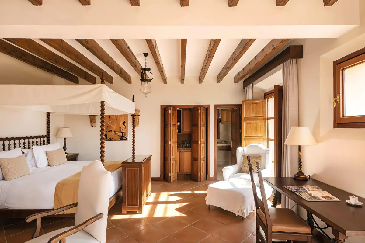 Sun-filled hotel bedroom with exposed beam ceiling, ornate wooden furnishings, and archway entrance