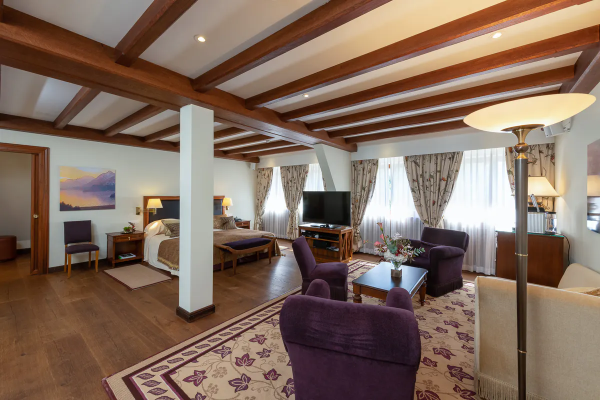 A junior suite with exposed wooden beams, purple armchairs, and corner windows overlooking the valley