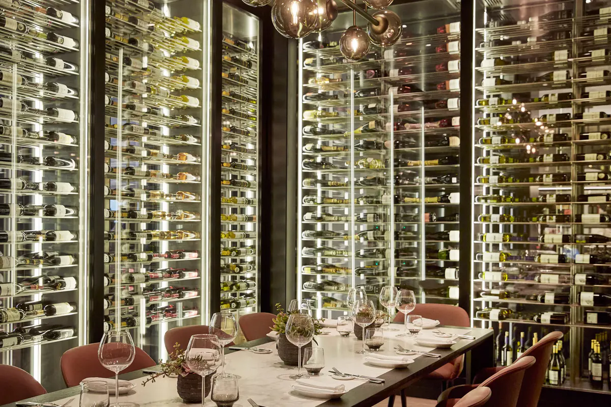 A private dining room with wine bottles wall-mounted behind glass, white tablecloths, and formal service