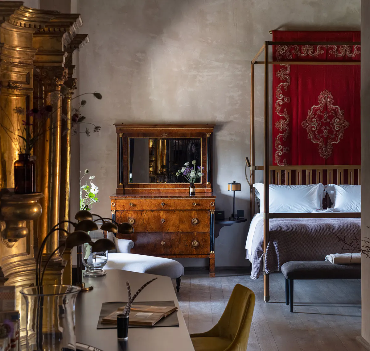 A suite bedroom with a red canopy bed, antique gilt furniture, and a mustard chair on stone floors