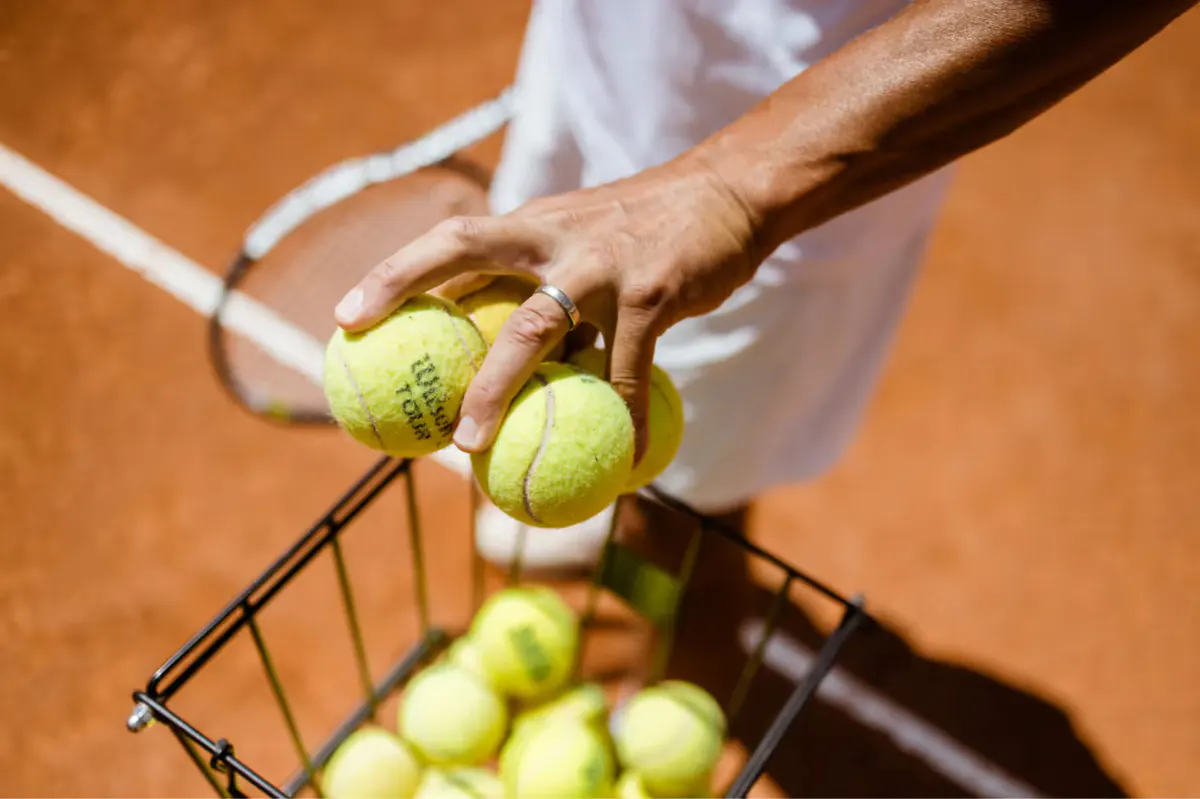 Tennis balls being held above a clay court with a racket and basket of additional balls