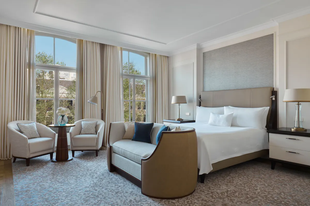 A room with a brown upholstered bed frame, cream seating, and tall arched windows overlooking the canal