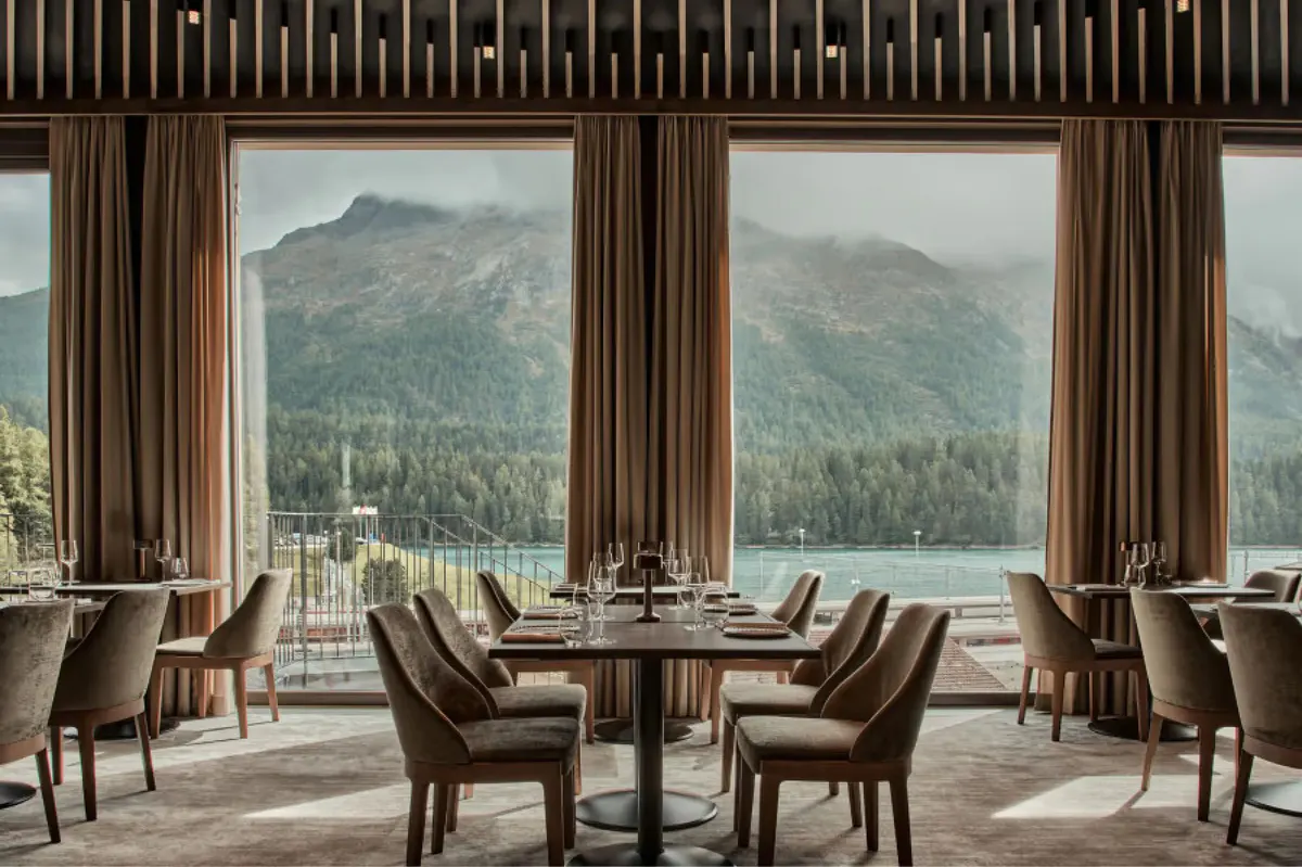 Restaurant with wood paneling, large windows framing alpine landscape, round dining tables