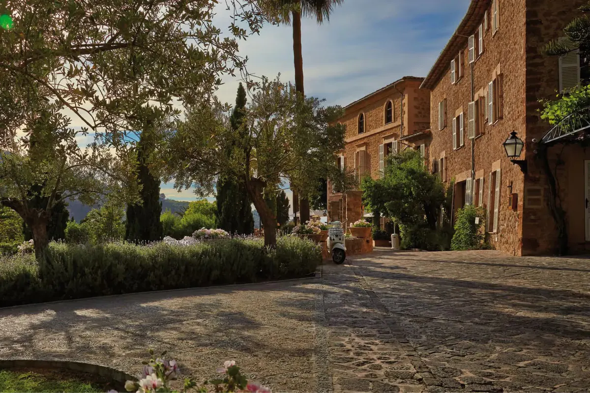 Stone pathway between rustic buildings lined with cypress trees and flowering plants