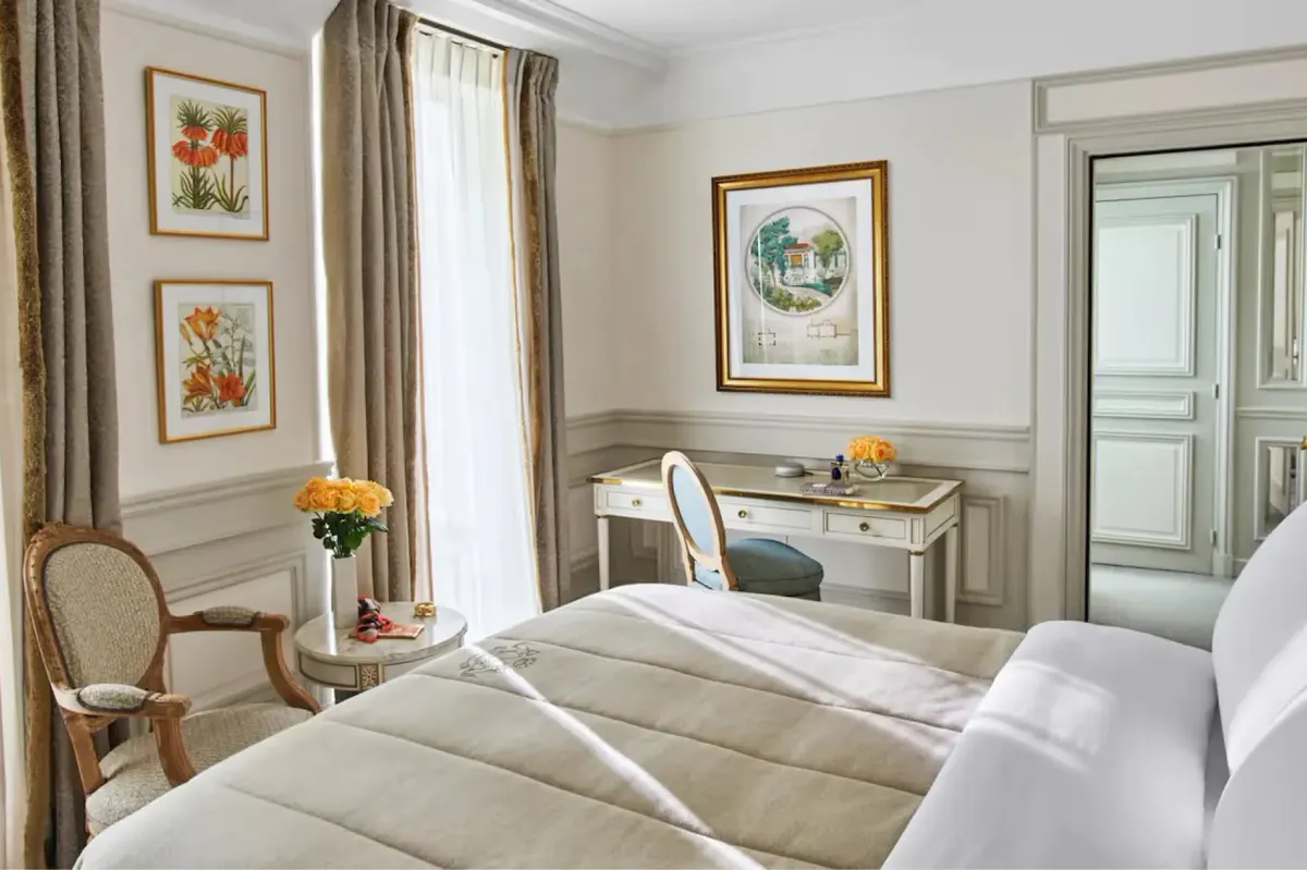 A suite with queen bed, pale furnishings, framed prints, and tall windows with views
