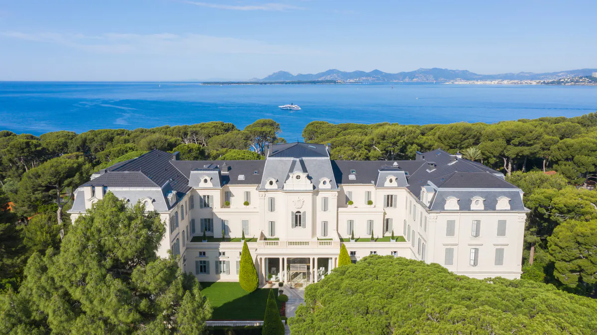 An aerial view of a cream-colored mansion with mansard roofs overlooking the Mediterranean and distant coastline