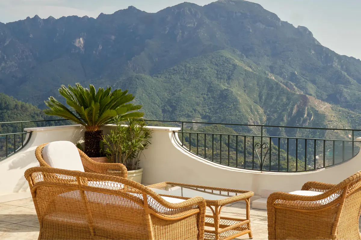 Terrace with wicker chairs, palm plants, turquoise Mediterranean vista and distant mountains at dusk
