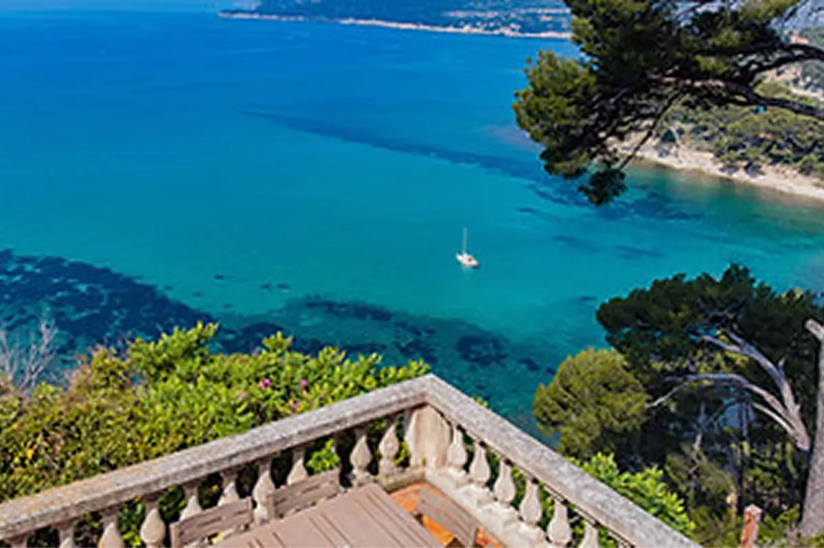 Terracotta terrace with stone balustrade overlooking turquoise Mediterranean, sailboat on cove, pine forest below
