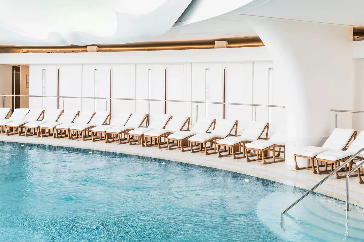 Turquoise indoor pool with white cushioned loungers, bright skylights, and bronze ceiling beams