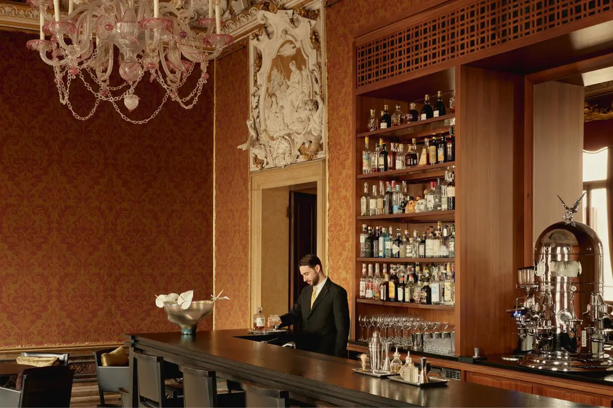 A classic bar with dark red walls, brass fixtures, a bartender, bottle-lined shelves, and crystal chandelier above