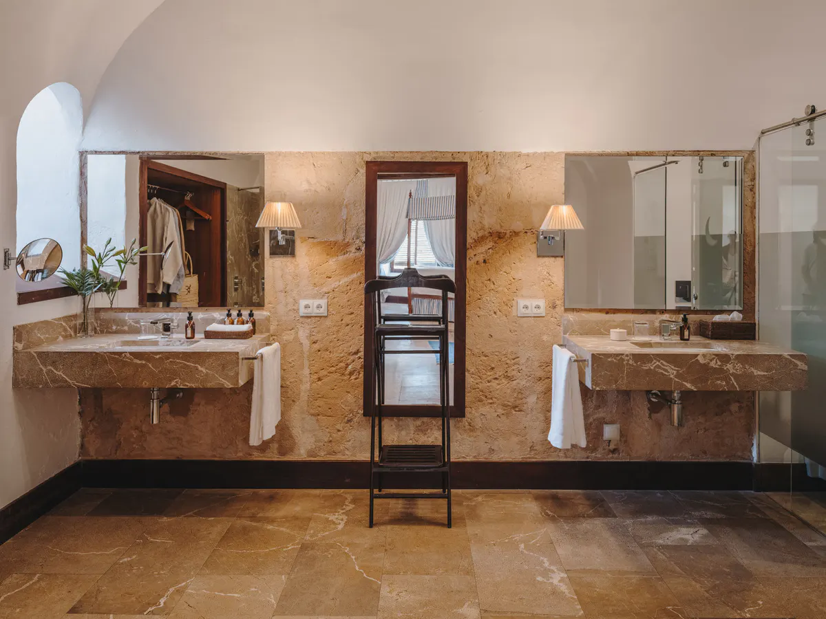 A dual vanity bathroom in warm stone with brass fixtures, mirrors, white towels, and pale archways