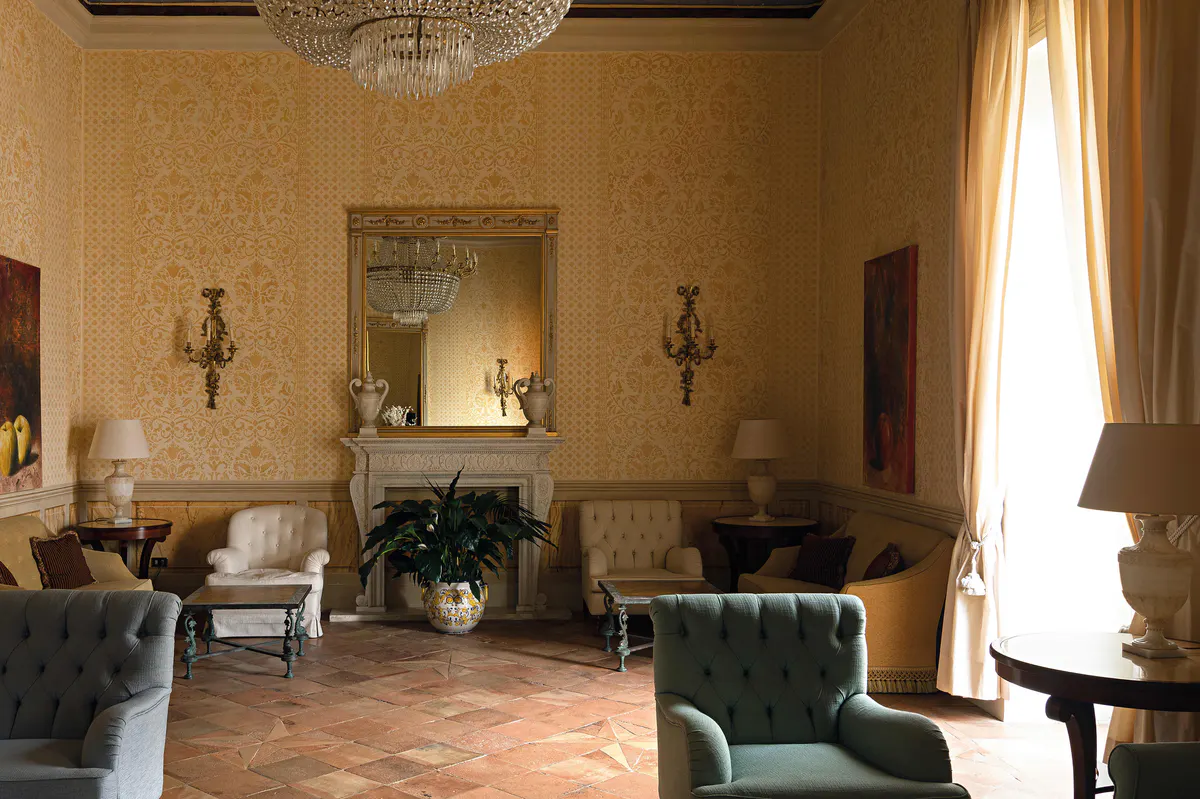 Grand salon with golden ochre walls, ornate ceiling fixture, fireplace, mirrors, potted plants, and lounge seating