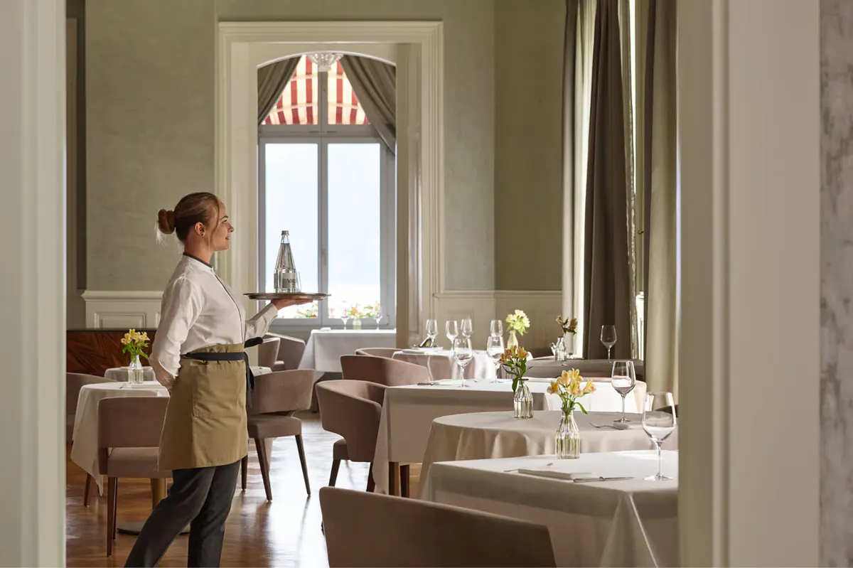 Private dining room with pale walls, server in white service dress, white-clothed tables, and tall windows