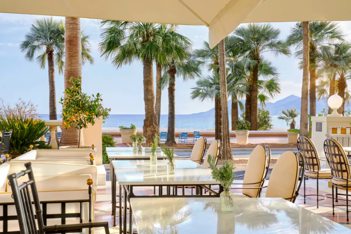 Beachside terrace dining with cream-colored umbrellas, palm trees, and sea views beyond
