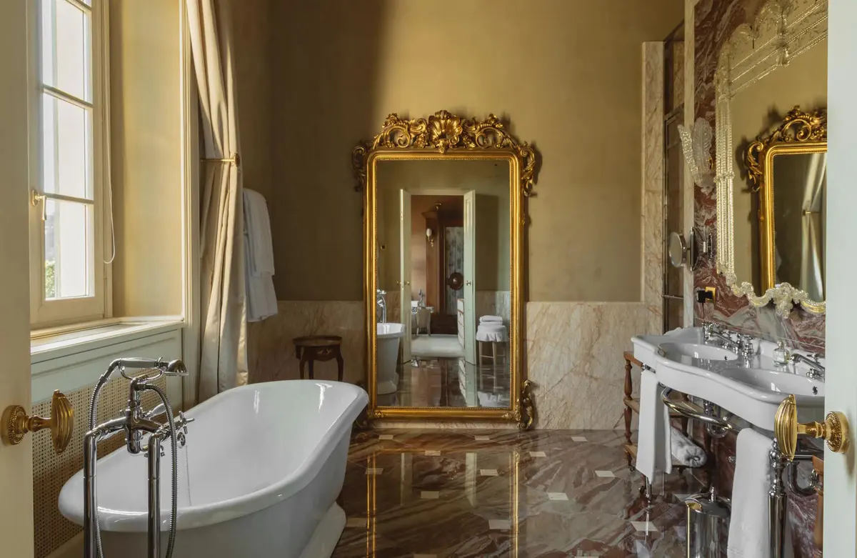 A pale bathroom with freestanding tub, gold-framed mirror, and marble wainscoting