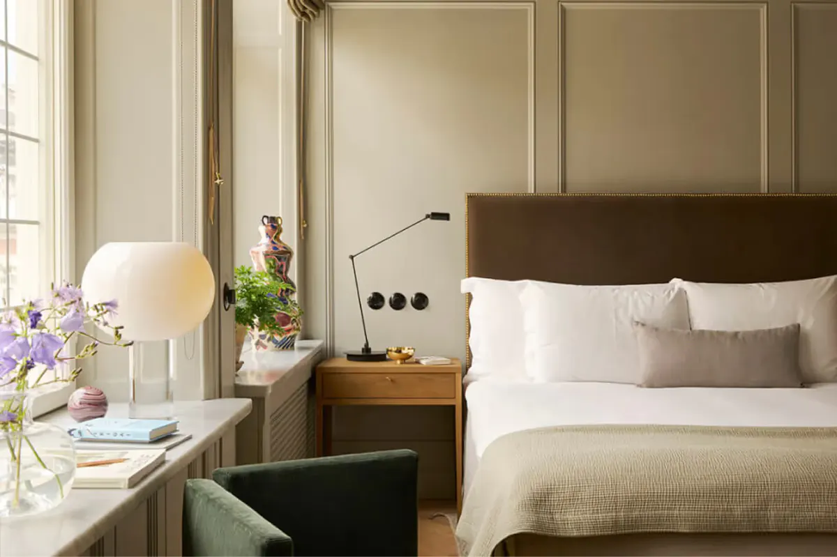 A bedroom with tan headboard, white pillows, glass bedside table, and flowers in a vase by the window