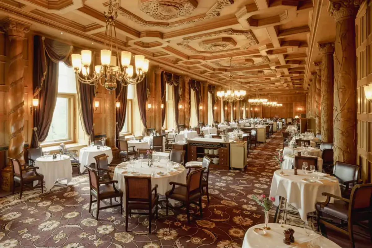 An elegant dining room with ornate gold ceiling, brass wall sconces, and white linens on every table