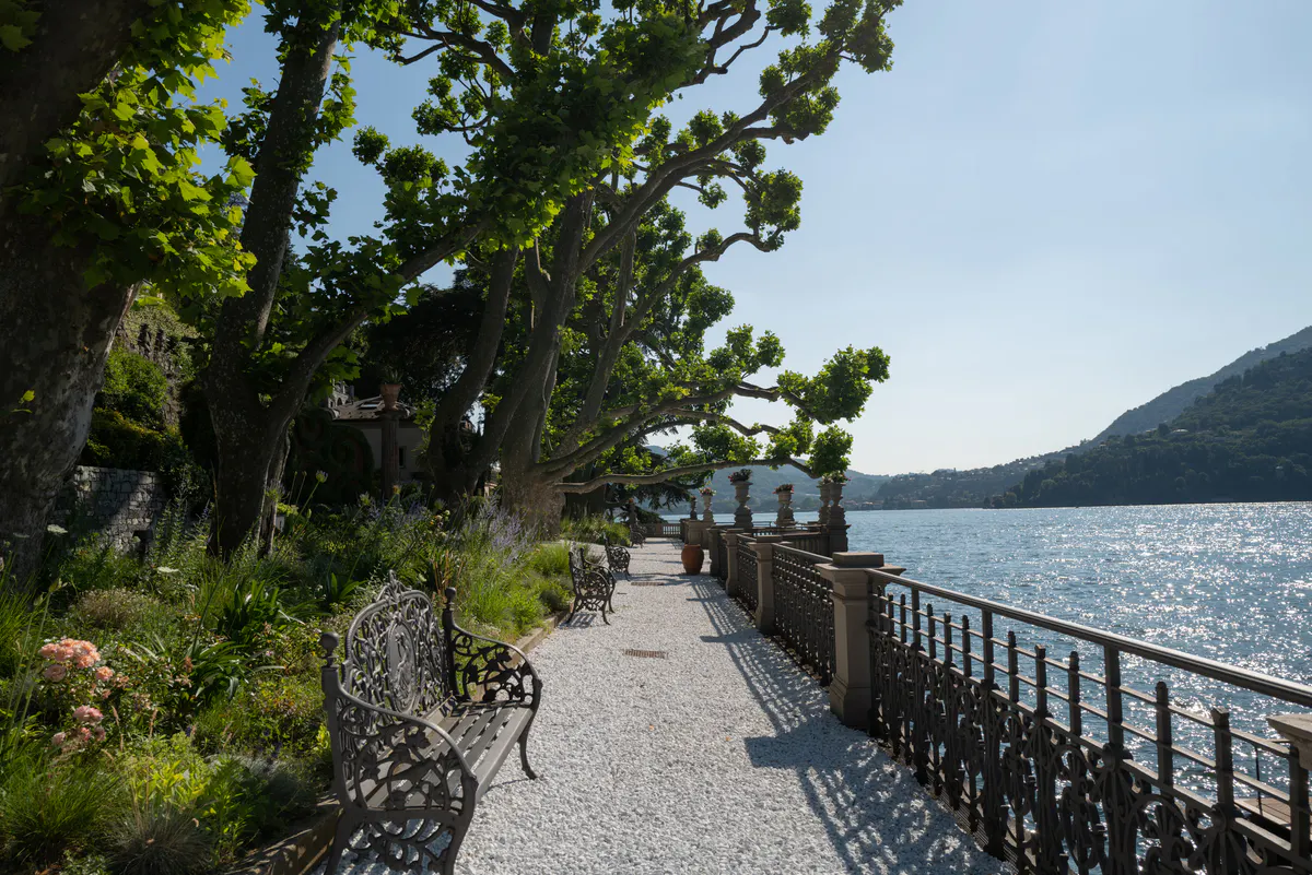 Waterfront pathway lined with mature trees, railing, and distant mountain peaks across the lake