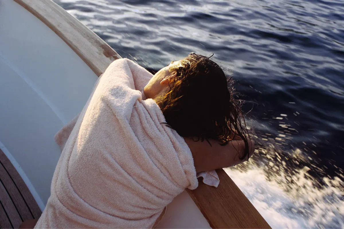 Person leaning over boat rail gazing at sea with coastal mountains in distance