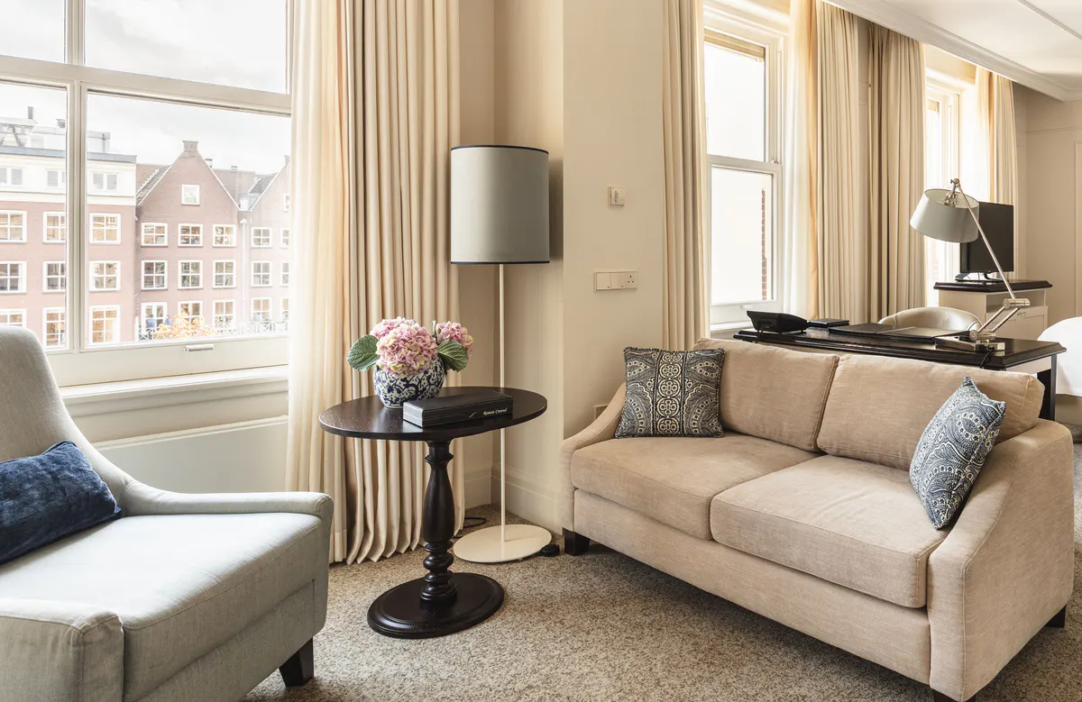 A sunlit lounge with cream armchairs and a beige sofa framing a view of 17th-century buildings across a canal