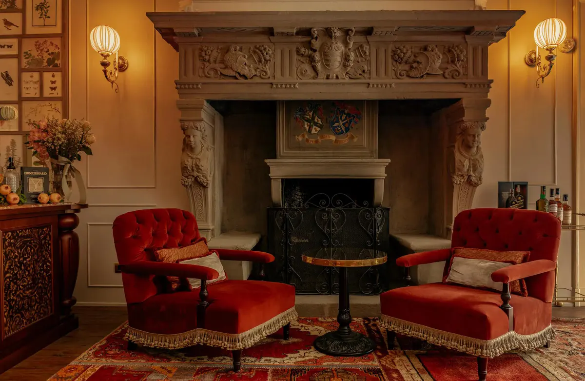 A fireplace with ornate carved mantel, red seating, and framed artwork