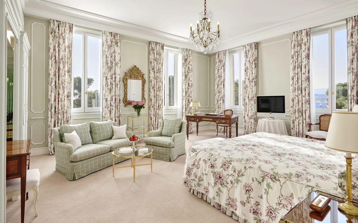 A spacious suite with tall windows, dual sitting areas, patterned drapery, and classical décor