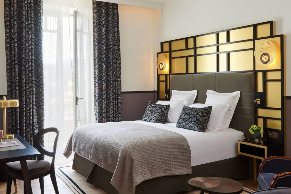 A bedroom with brass headboard and geometric patterned bedding, windows with coastal views