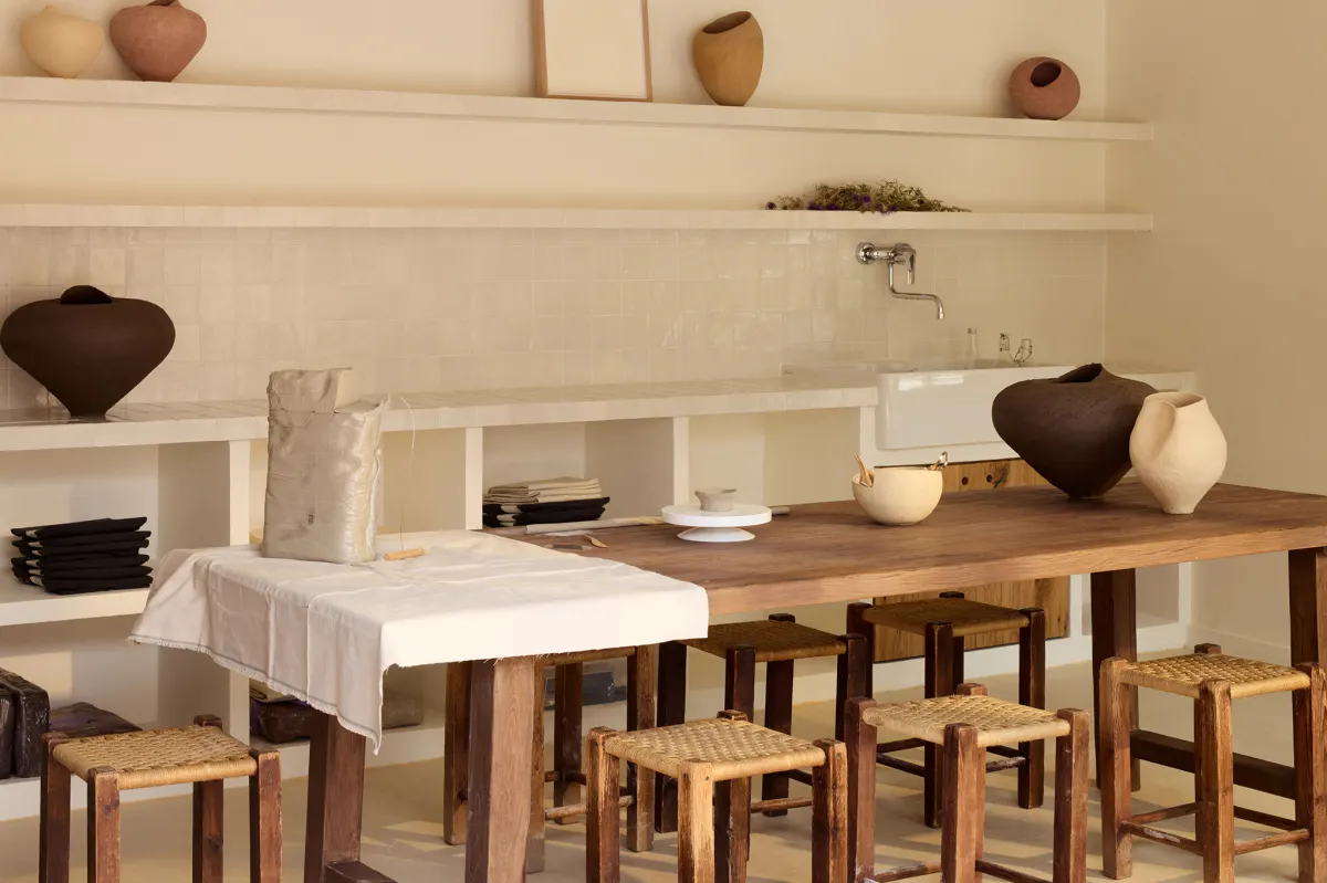 Dark and pale ceramic vessels line a wooden table beneath a tiled backsplash and shelf