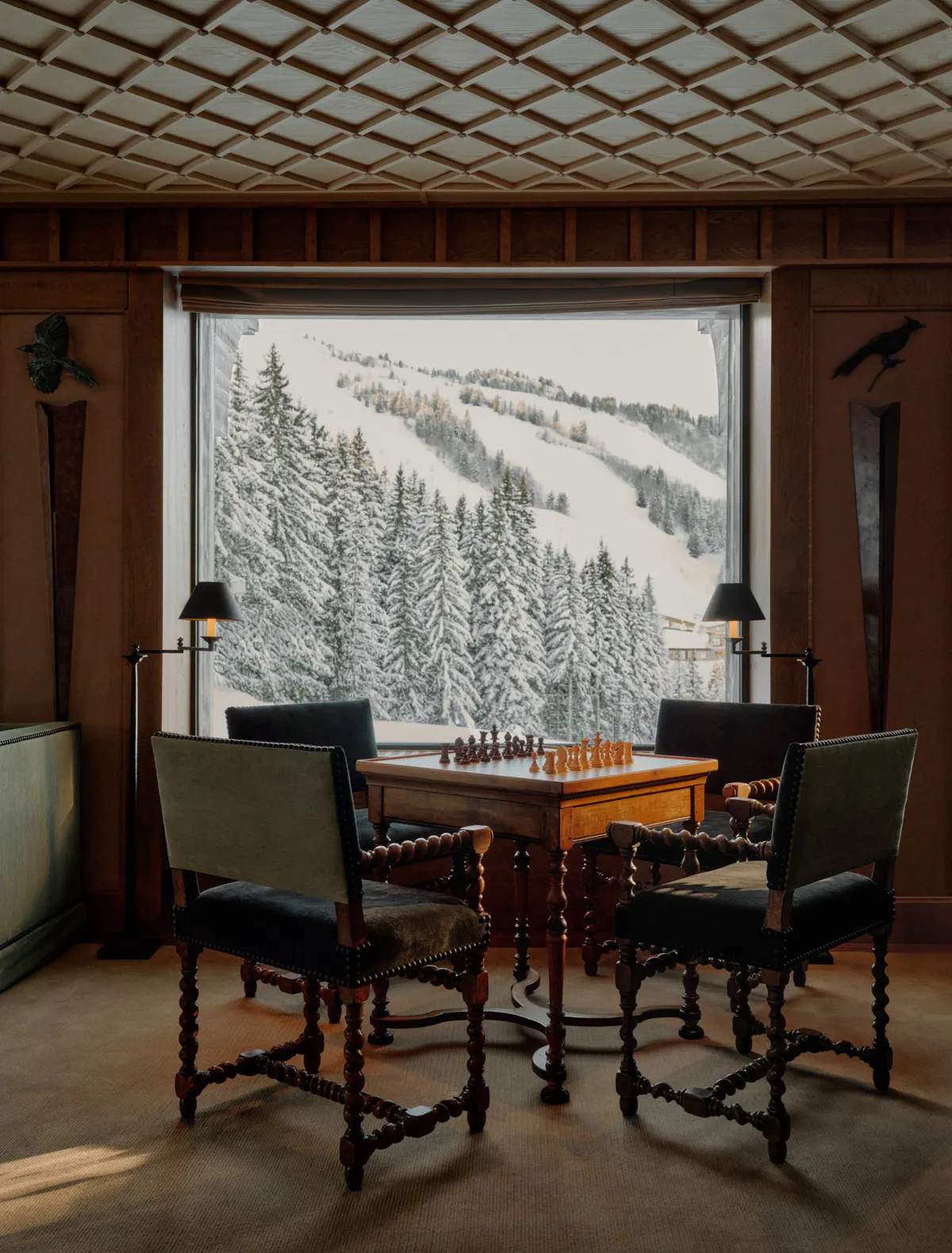 Chess table surrounded by four chairs beneath a coffered ceiling, facing a window over the piste