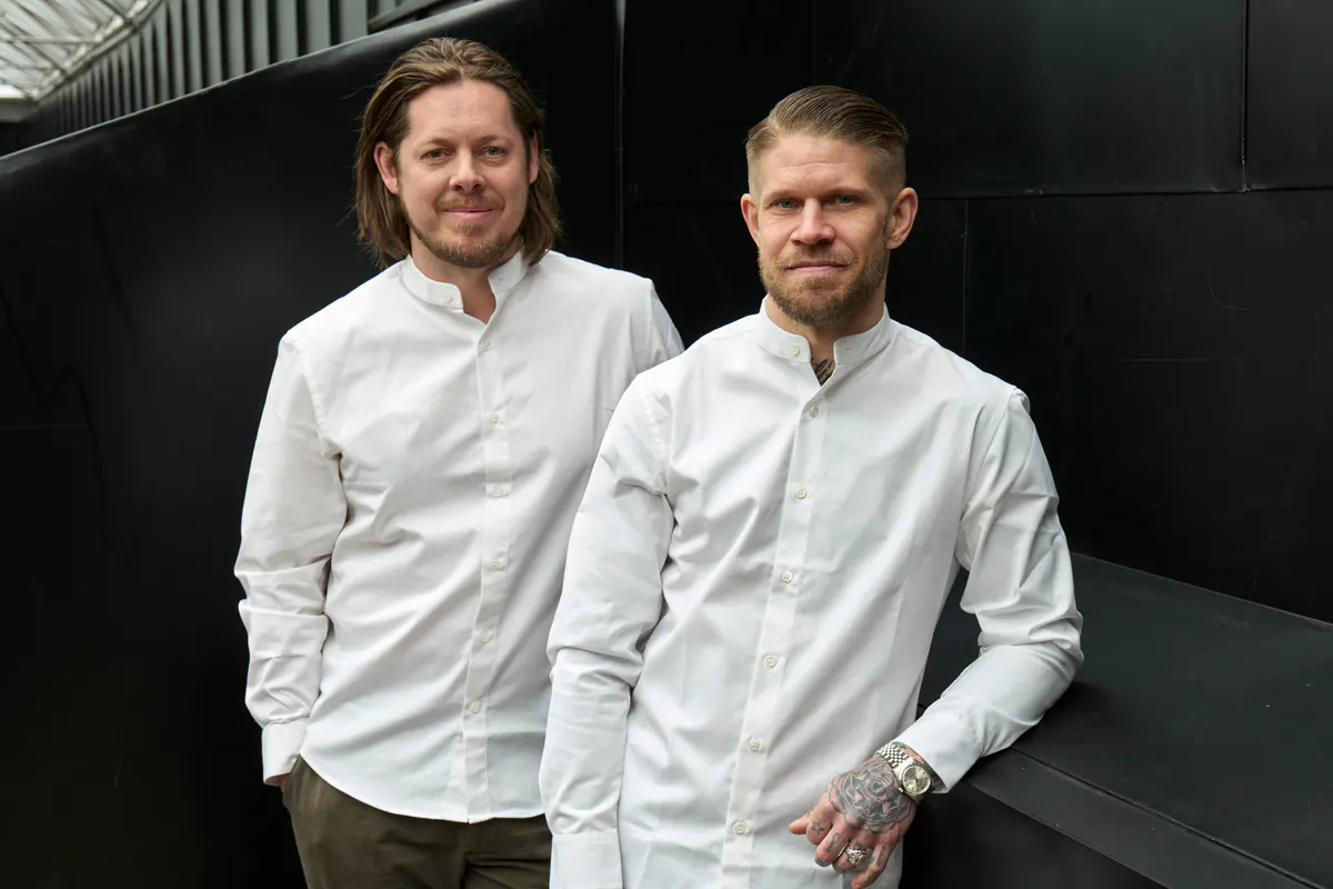 Two chefs in white chef coats standing before their restaurant kitchen