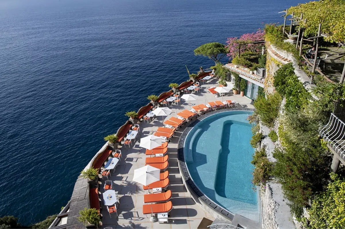 Infinity pool terrace lined with orange loungers, white umbrellas, ivy-clad stone, turquoise Mediterranean below