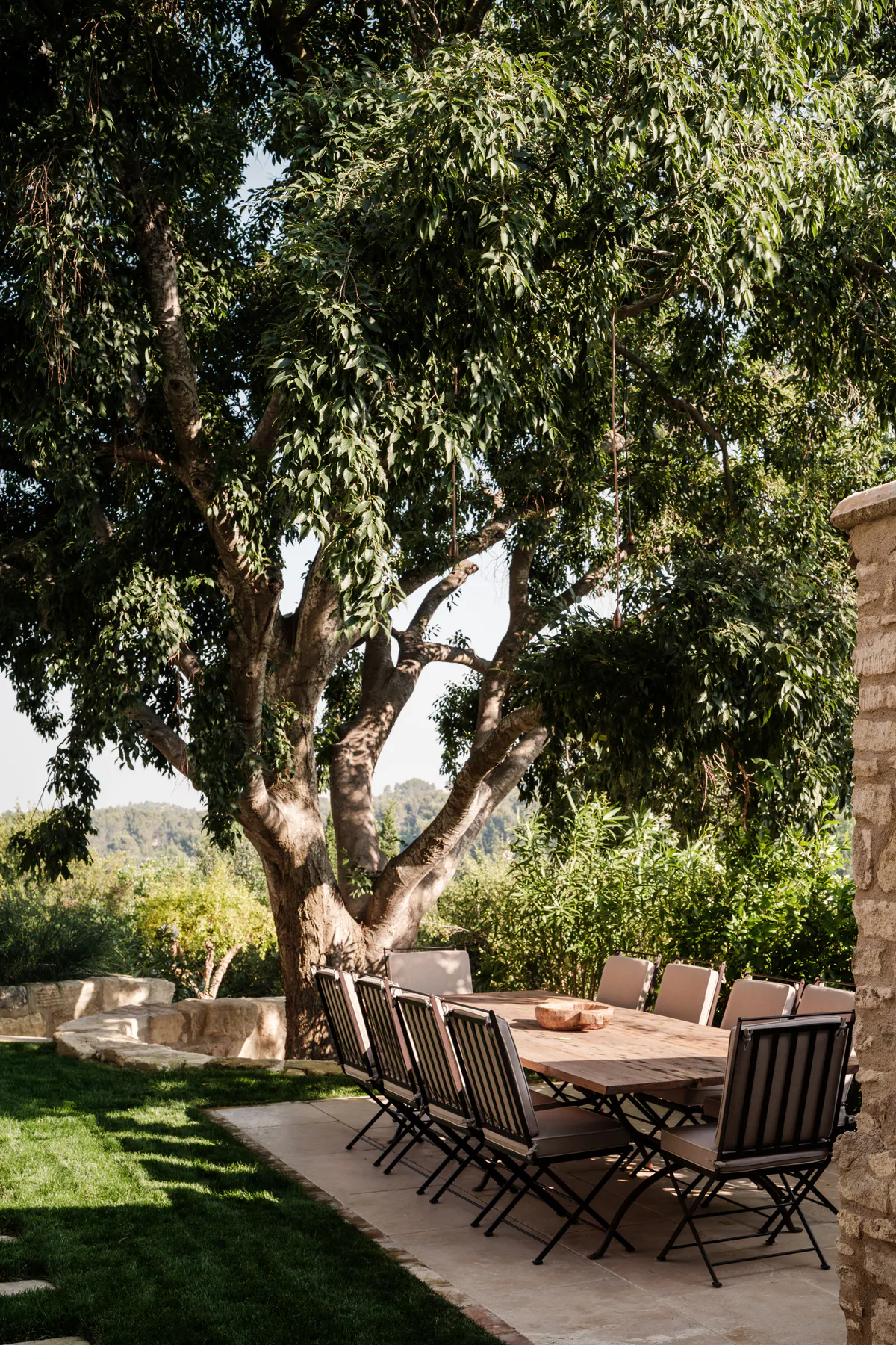 Stone villa terrace beneath mature olive tree with wooden table, cypress borders, mountain landscape