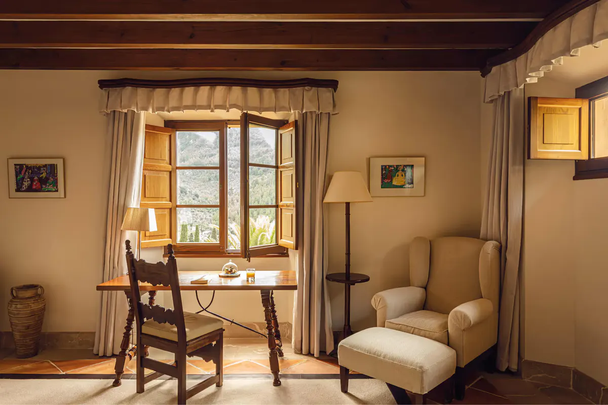 Suite with beamed wooden ceiling, window desk, upholstered chair, and warm cream walls
