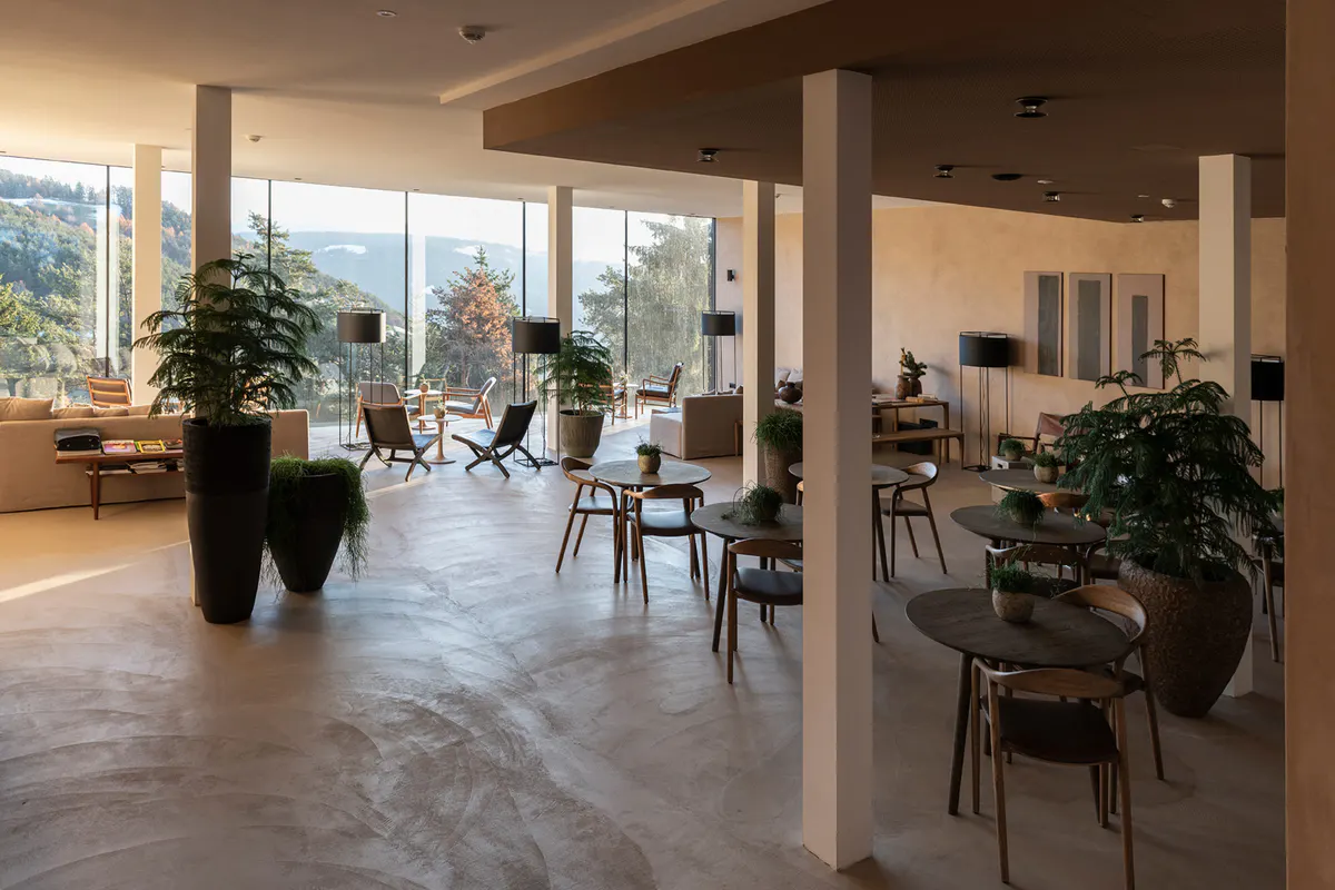 Large dining hall with concrete floor, tall support columns, and floor-to-ceiling windows overlooking mountains