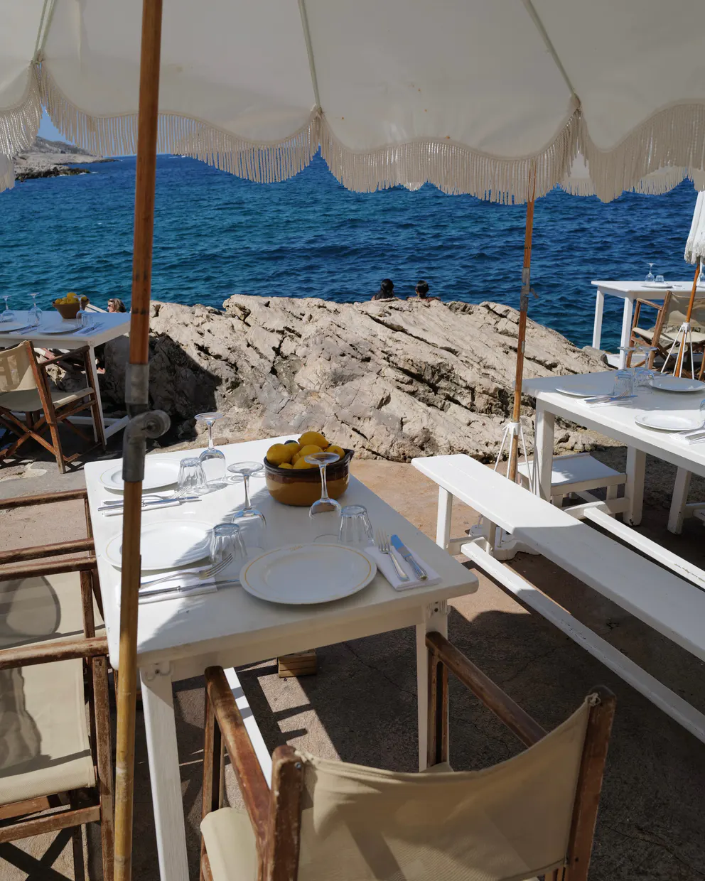 A set table under a fringed parasol at Tuba Club, the rocks and open sea beyond