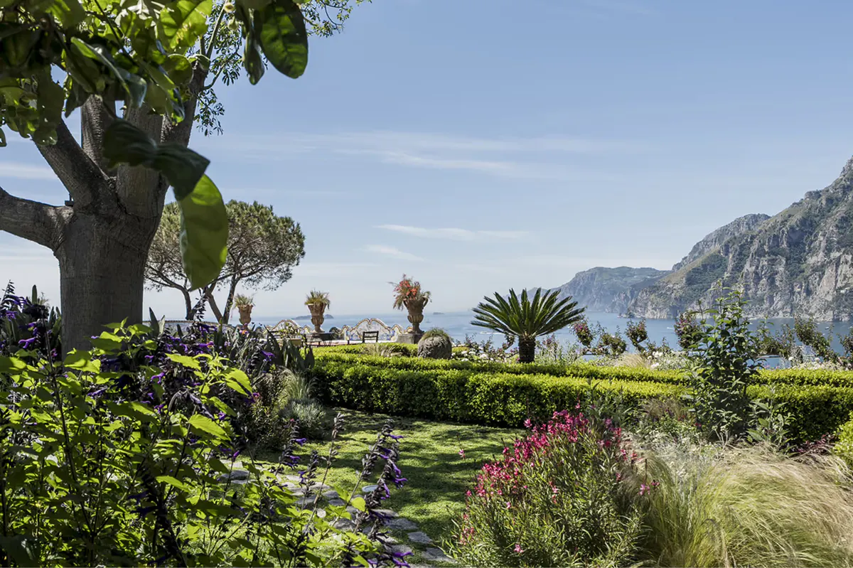 Landscaped garden with flowering shrubs, mature trees, palms, and dramatic cliffside coastline in distance