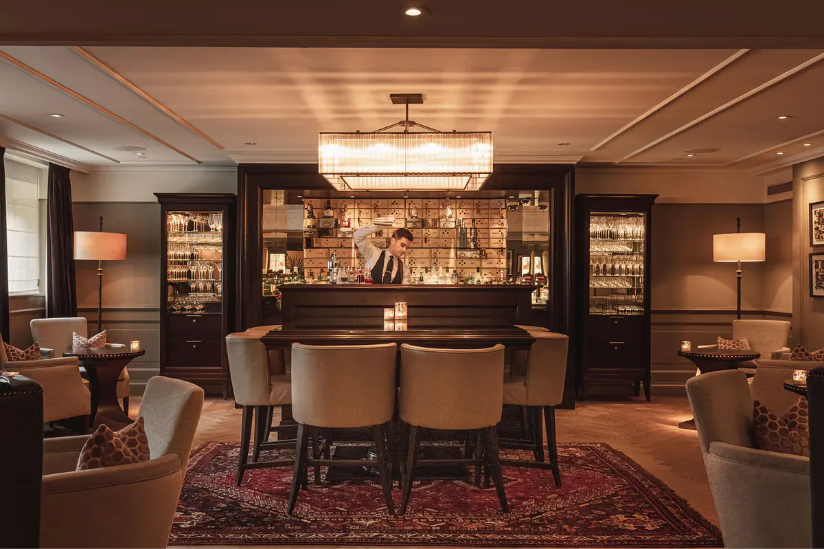 A bar lounge with dark woodwork, illuminated shelving displaying spirits, and upholstered lounge seating